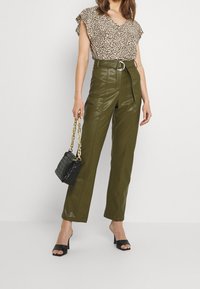 Top marrón de estampado floral con mangas rizadas, pantalones de cuero ecológico verdes de tiro alto, bolso negro trenzado y zapatos de tacón negros.