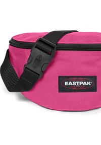 Růžový látkový ledvinka s černým nastavitelným popruhem a sponou. Obsahuje logo Eastpak na černém štítku, zapínání na zip a zaoblený tvar.
