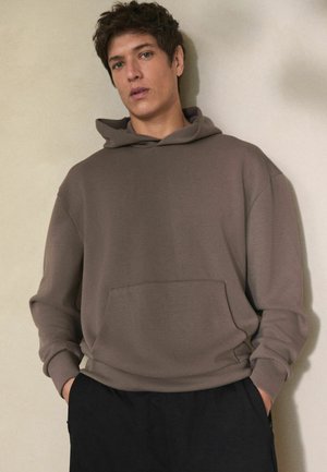 Brown hoodie with a front pocket, relaxed fit, adjustable drawstring hood, and ribbed cuffs, paired with black trousers. Simple design.