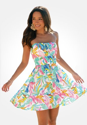 Kobieta uśmiechająca się, ubrana w kolorową, kwiecistą sukienkę na ramiączkach, kręcąca się w sukience, z długimi ciemnymi włosami i kolczykami w formie frędzli.