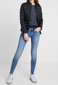 Svart quiltad jacka ovanpå en marinblå och vit randig skjorta, tillsammans med ljusblå slitna slim fit-jeans och vita sneakers.