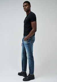 Camiseta negra de manga corta, jeans azules desgastados con detalles rasgados, y botas negras con cordones. Pose de pie con las manos en los bolsillos.