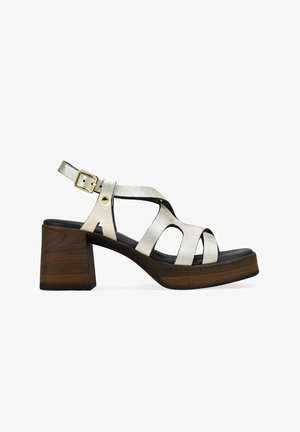 Sandalia blanca de cuero con tiras, hebilla dorada, tacón grueso de bloque de madera oscura y plantilla acolchada negra, mostrada de perfil izquierdo.