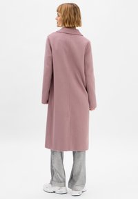Lungo cappotto rosa realizzato in tessuto texturizzato con colletto a contrasto e taglio dritto. Il modello indossa pantaloni argentati a zampa d'elefante e sneakers bianche.