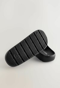 Czarne buty wsuwane z teksturowaną gumową podeszwą z poziomymi rowkami i okrągłymi akcentami. Wnętrze ma gładkie wykończenie.