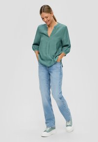 Tijlgroene, losvallende blouse met een V-hals en plooidetail, gecombineerd met lichtblauwe rechte jeans en groene sneakers met dikke witte zolen.