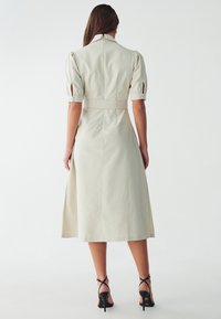 Vestido midi de color beige claro con un corsé ajustado, mangas abullonadas y una cintura ceñida con cinturón. Presenta un cuello y una abertura lateral para un detalle adicional.