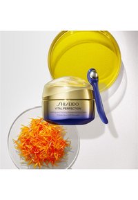 Shiseido Vital Perfection ögonkräm, blå och gul burk, tillsammans med en silverapplikator och orange blomsterdetaljer mot en vit bakgrund.