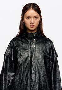 Mujer joven con cabello oscuro y lacio que lleva una chaqueta de cuero negra oversized con un alto cuello y botones de presión metálicos, mirando de frente.