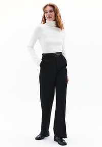 Weißer Rollkragenpullover kombiniert mit hochgeschnittenen schwarzen Hosen. Die Hosen verfügen über einen Gürtel mit einer goldenen Schnalle. Glatte Stofftextur.