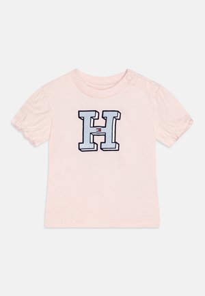 Camiseta de algodón rosa con mangas abullonadas; presenta una gran letra "H" bordada en azul marino y blanco con un pequeño acento rojo.