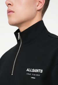 Férfi fekete cipzáras felsőt visel, amelyen az "ALLSAINTS" és japán szöveg hímzett a nyak alatt, a bal mellen.