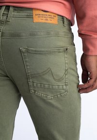 Jeans de mezclilla verde oliva con un bolsillo trasero que presenta un diseño de costura curvada y una etiqueta de cuero marrón encima con el nombre de la marca y un lema.