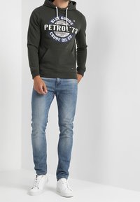 Sudadera con capucha verde oscuro con cordones blancos y bolsillo frontal, con un gran gráfico impreso en azul y blanco. Combinada con vaqueros azul claro y zapatillas blancas.