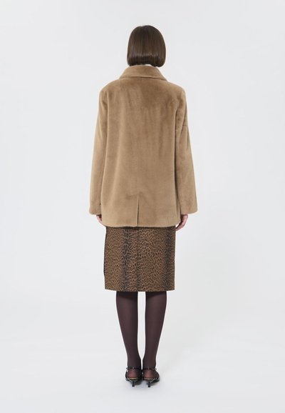 Cappotto in ecopelliccia marrone con un ampio colletto, abbinato a una gonna a tubino con stampa leopardata. Calze scure e tacchi completano l'outfit.