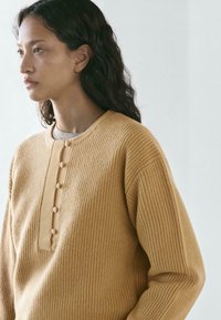 Maglione giallo senape a coste con scollo rotondo e patta con cinque bottoni. Tessuto morbido e strutturato, vestibilità comoda.