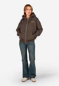 Barna zipzáros kapucnis pulcsi nagy elülső zsebekkel, húzózsinórral ellátott kapuccsal és a bal mellen hímzett "PEGADOR" felirattal. Kék trapéz szabású farmernadrággal viselve.