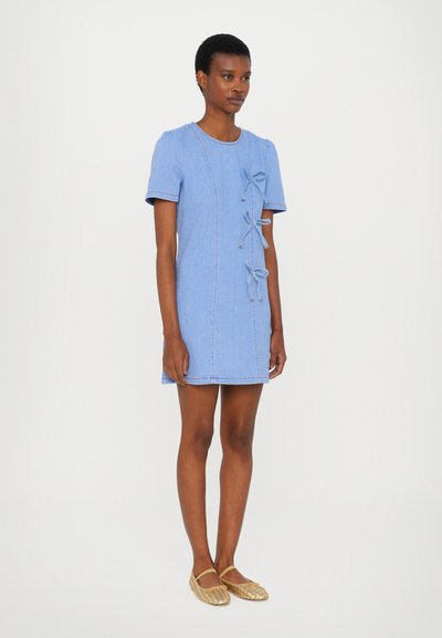Light blue denim dress with short sleeves, decorative bows on the side, and visible stitching details. Worn with woven, flat shoes.