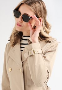 Ochelari de soare negri cu accesorii aurii, formă rectangulară, purtați de o persoană în trench de culoare bej deschis peste un top cu dungi, pe un fundal simplu.