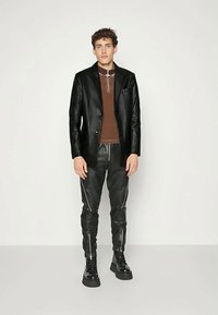 Juodas tekstūruotas bleizeris ant rudos džemperio, derinamas su juodomis odinėmis kelnėmis su užtrauktukų akcentais ir juodais masyviais sportbačiais.