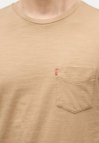 Κοντινό πλάνο ενός μπεζ T-shirt με στρογγυλή λαιμόκοψη και θήκη στο στήθος που διαθέτει μια μικρή κόκκινη ετικέτα Levi’s στην αριστερή πλευρά.