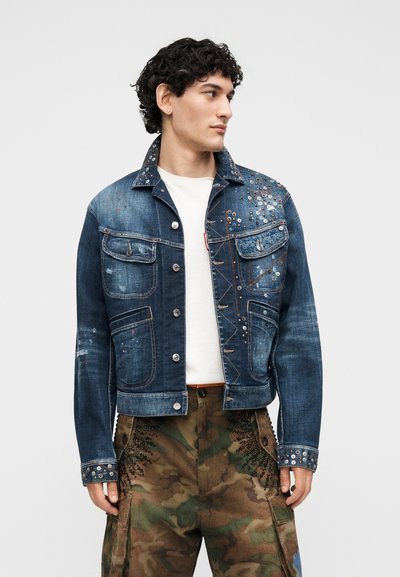 Άνδρας που φοράει μπλε τζιν μπουφάν με τρουκς πάνω από λευκό πουκάμισο, συνδυασμένο με παντελόνι παραλλαγής διακοσμημένο με μαύρα χάντρες.