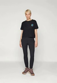 Svart avkortet t-skjorte med et fargerikt sirkulært logo på venstre bryst, kombinert med smale svarte bukser og brune treningssko.