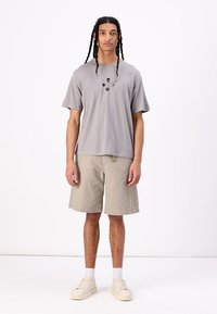 Ljusgrå bomulls-T-shirt med kort ärm och svarta grafiska detaljer, kombinerad med beige knälånga shorts och krämfärgade sneakers.