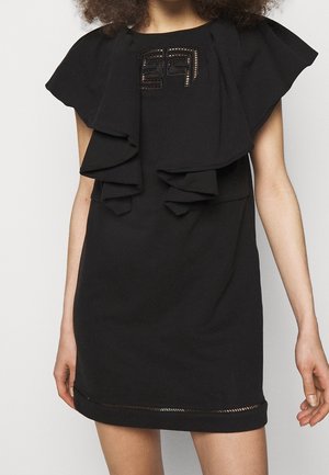 Femme portant une robe noire sans manches avec un détail de volants superposés sur la poitrine et un patch brodé subtil près de l'encolure.