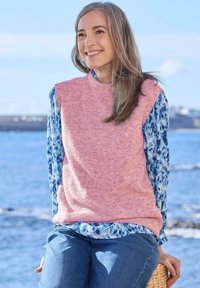 Eine Frau lächelt am Meer, trägt einen rosa ärmellosen Pullover über einer blauen gemusterten Bluse und blauen Jeans und sitzt auf einem geflochtenen Hocker.