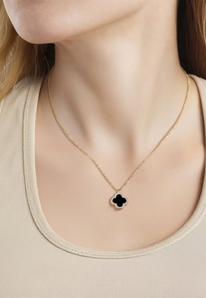Goldene Halskette mit schwarzem vierblättrigem Kleeblatt-Anhänger, umrandet von kleinen Diamanten, an einer Person mit beigem Oberteil.