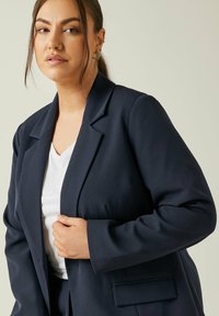 Granatowa marynarka z klapami i dwiema przednimi kieszeniami, zestawiona z białym topem w serek. Gładki materiał o dopasowanym kroju.