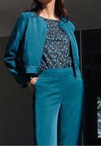 Teal cropped jacket over a patterned blouse with blue, black, and gray accents; fitted pants in matching teal; smooth fabric texture.