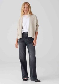 Cardigan beige a trama sopra una t-shirt bianca, abbinato a jeans grigi scuri a vita alta. Il modello indossa scarpe basse nere.