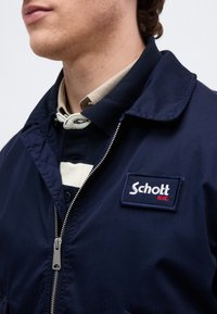 Jeune personne portant une veste zippée bleu marine avec un écusson du logo Schott N.Y.C. sur une chemise à col rayée beige et bleu marine.