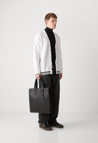 Cárdigan blanco con textura acanalada, suéter de cuello alto negro, bolso tote negro, pantalones de mezclilla negros y zapatillas oscuras, de pie frente a un fondo liso.