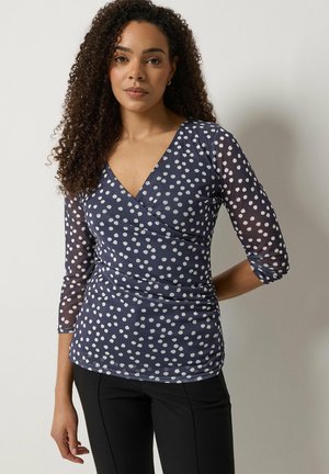 Vrouw met krullend haar, gekleed in een marineblauw wikkelshirt met witte polka dots en driekwart doorschijnende mouwen, en zwarte broek, staand tegen een effen muur.