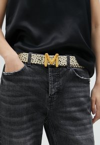 Ceinture crème à pois noirs, ornée d'une boucle en forme de lettre "M" dorée et texturée. Portée avec un jean gris foncé déchiré.