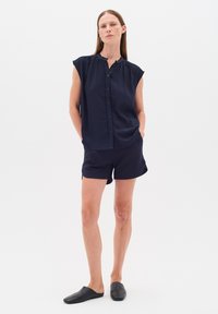 Mörkblå ärmlös skjorta med knäppning och rynkad rundad halsringning, matchad med shorts i samma färg. Modellen bär svarta slipskor.