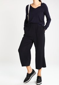 Woman wearing a navy V-neck sweater, loose black cropped pants, and black platform oxford shoes, standing with hands in pockets.