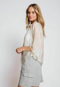 Light, sheer blouse in a muted cream color with a subtle pattern, featuring a button-up front and wide sleeves, paired with gray cargo shorts.