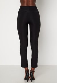 Pantalon noir ajusté avec une texture lisse, présentant une taille mi-haute, des poches arrière et une coupe fuselée. Porté avec des escarpins noirs.