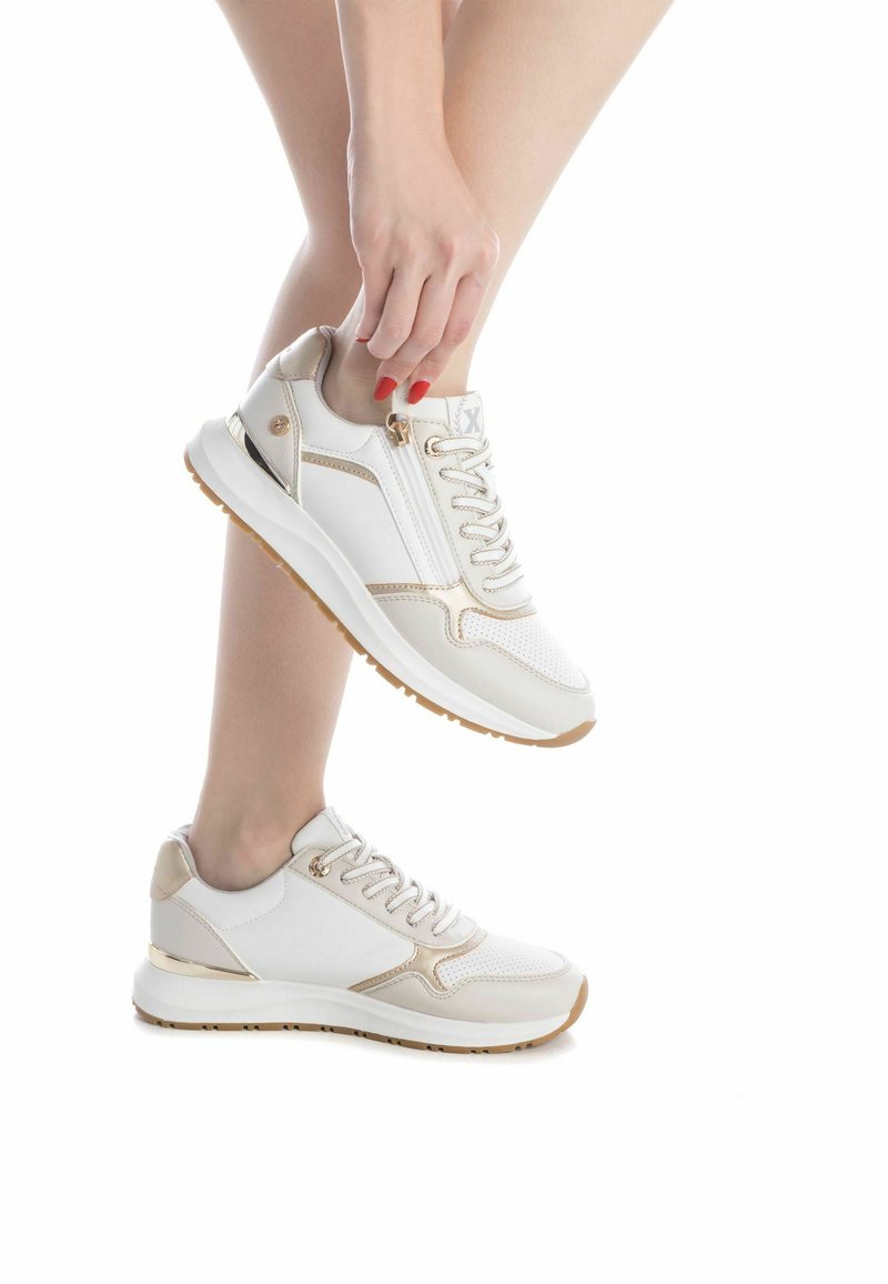 Zapatillas deportivas blancas y doradas con diseño de cordones, detalles perforados y tobillo acolchado, con una suela de goma resistente.