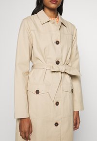 Femme portant un trench-coat beige ceinturé avec boutons sur le devant et poches à rabat, par-dessus un haut à carreaux orange, debout devant un fond uni.