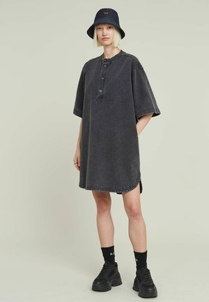 Robe oversize en denim gris avec des manches courtes, boutonnage sur le devant, ourlet arrondi et coutures contrastées. Portée avec un chapeau seau bleu marine et des baskets noires.