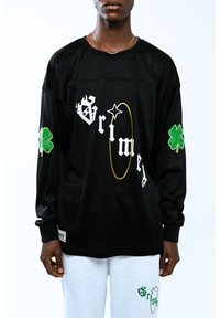 Jersey negro de manga larga con tejido de malla. Presenta texto blanco, tréboles bordados en verde y acentos dorados. Llevado con pantalones de chándal grises.