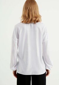 Langærmet hvid bluse med en løstsiddende pasform, blød tekstur, let samlede ærmer og rund halsudskæring. Flad kant med bløde draperinger.
