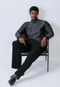 Czarna koszula w wzory z kołnierzykiem, wykonana z gładkiego materiału. Połączona z luźnymi czarnymi spodniami i błyszczącymi czarnymi butami.