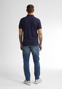 Námořnická polo tričko s krátkým rukávem, spárované se světle modrými džíny. Viditelný detail tetování na paži, s bílými teniskami a hladkým povrchem.