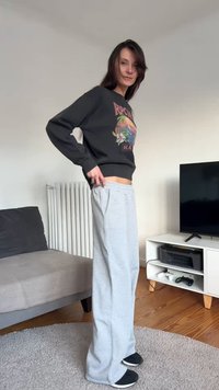 Sive trenirke z bočnimi žepi, kombinirane s črno majico z okroglim izrezom in barvnim grafičnim vzorcem. Črni čevlji brez vezalk. Prostoren kroj.